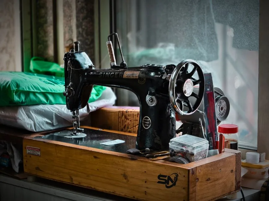 A treadle sewing machine sits near a window in a room with wooden floors.
