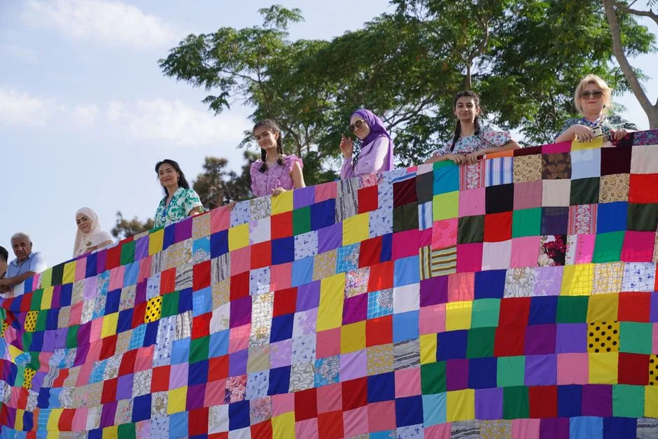 Two volunteers hold up a large, colorful quilt for an audience to see.