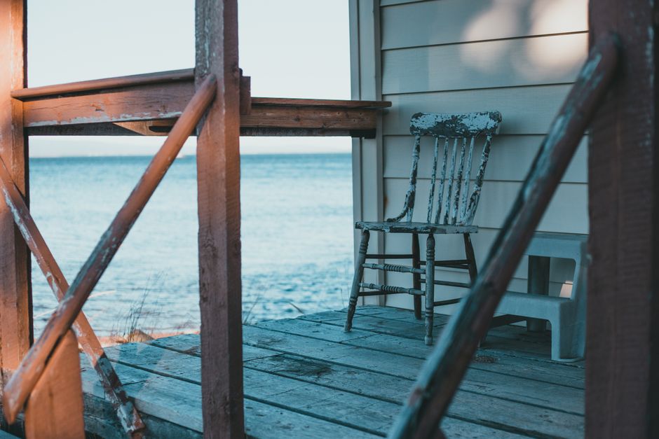 An empty chair on a porch faces the water as the sun sets.